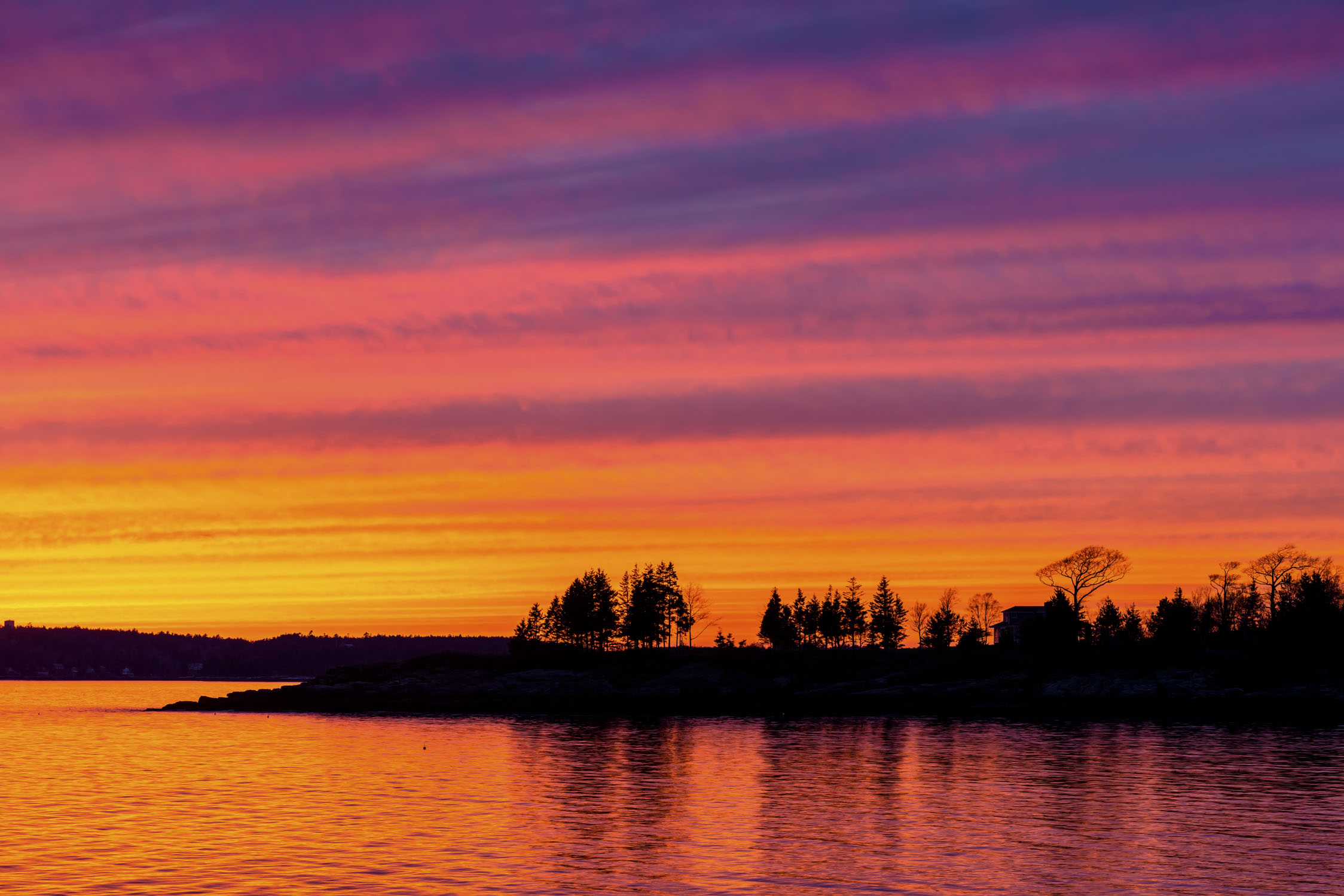 Water in the foreground reflects the sky, then there is a strip of land with silouhettes of trees and a house, with a bands of color across the sky, starting with blue then purple then orange then yellow.