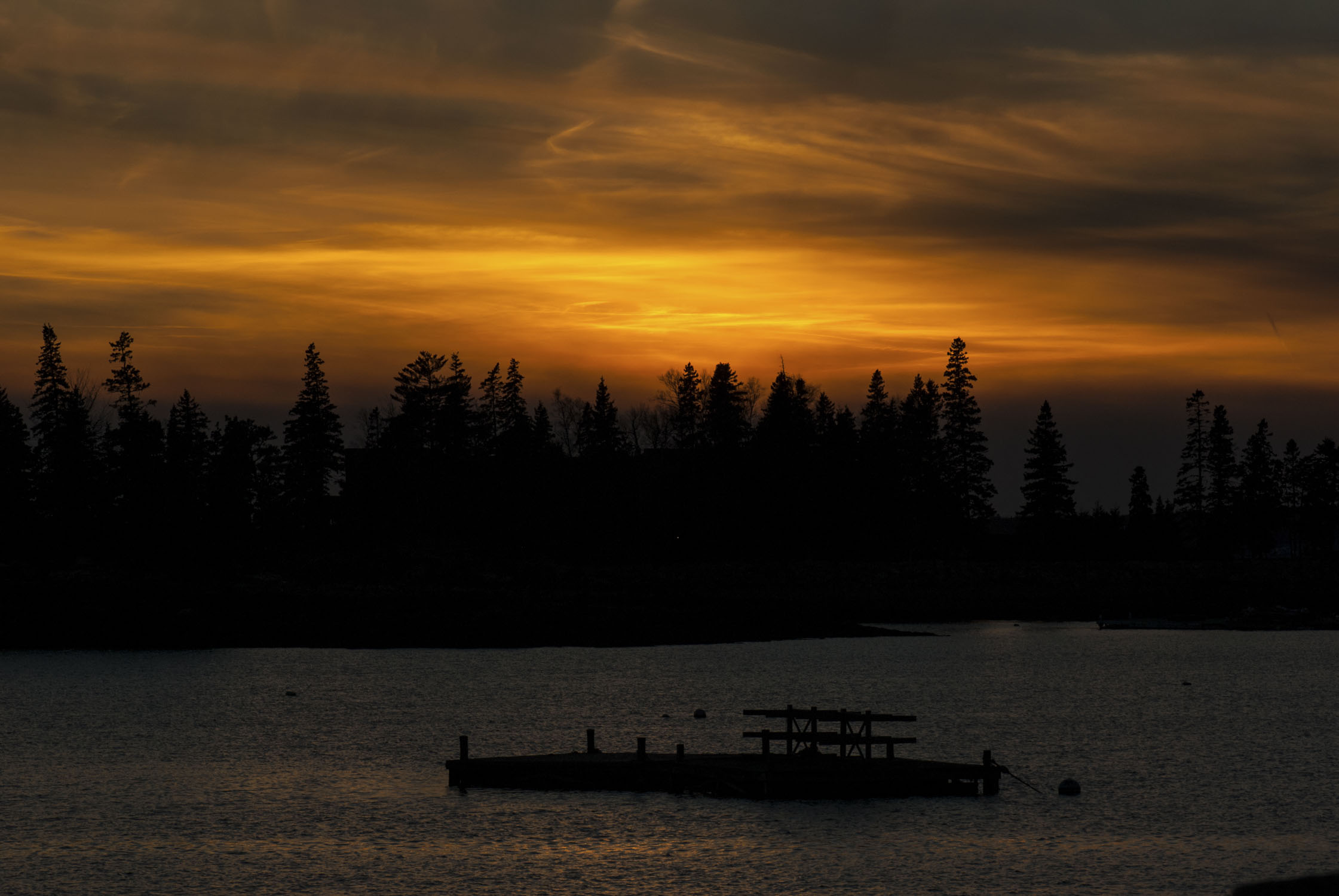 A late dusk sky with yellow and black at the top, a band of trees in the middle, and water with a float at the bottom.