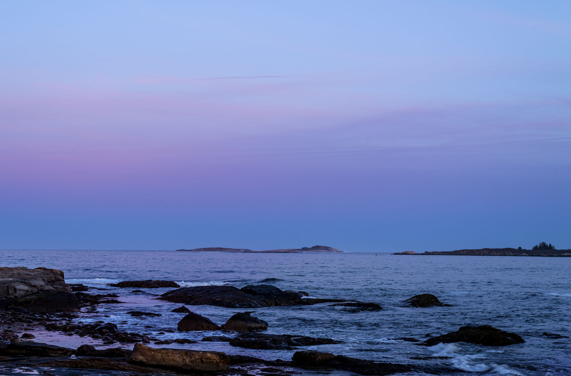 The sky is pink and blue in the twop two thirds, and the bottom is ocean with rocks.