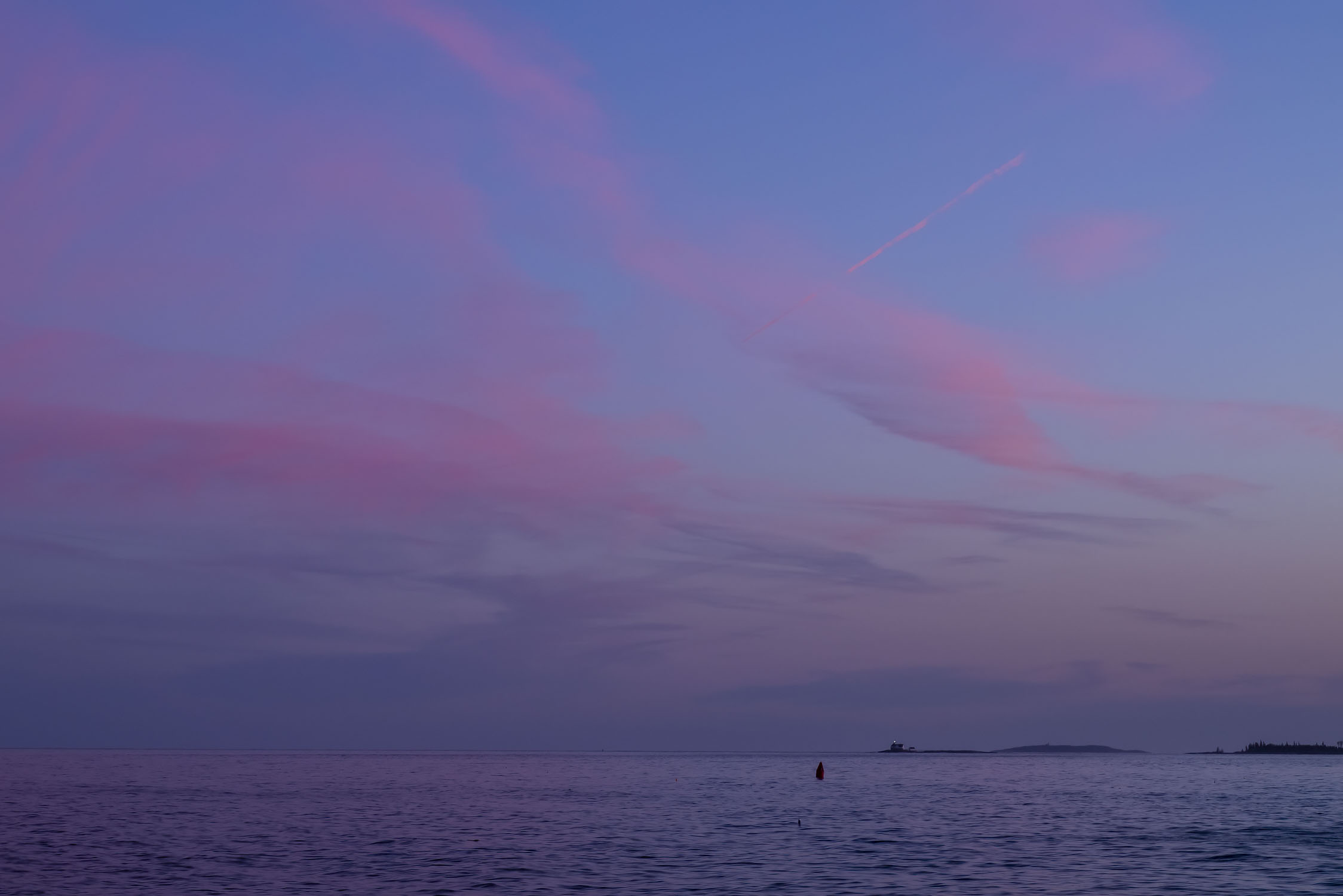 Most of the photo shows the sky in blue and pink, while the very bottom has water with a bouy and a distant lighthouse