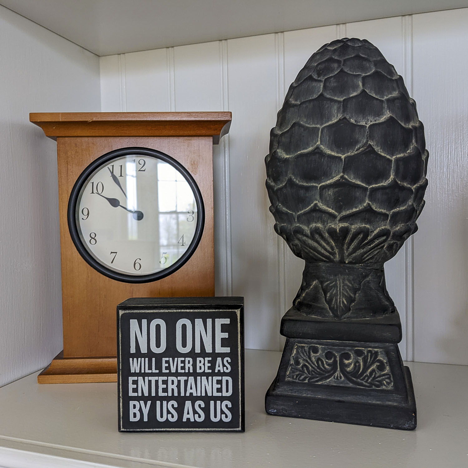A display nook that has a clock, a sculpture of a pine cone, and a sign that states No one will ever be as entertained by us as us.