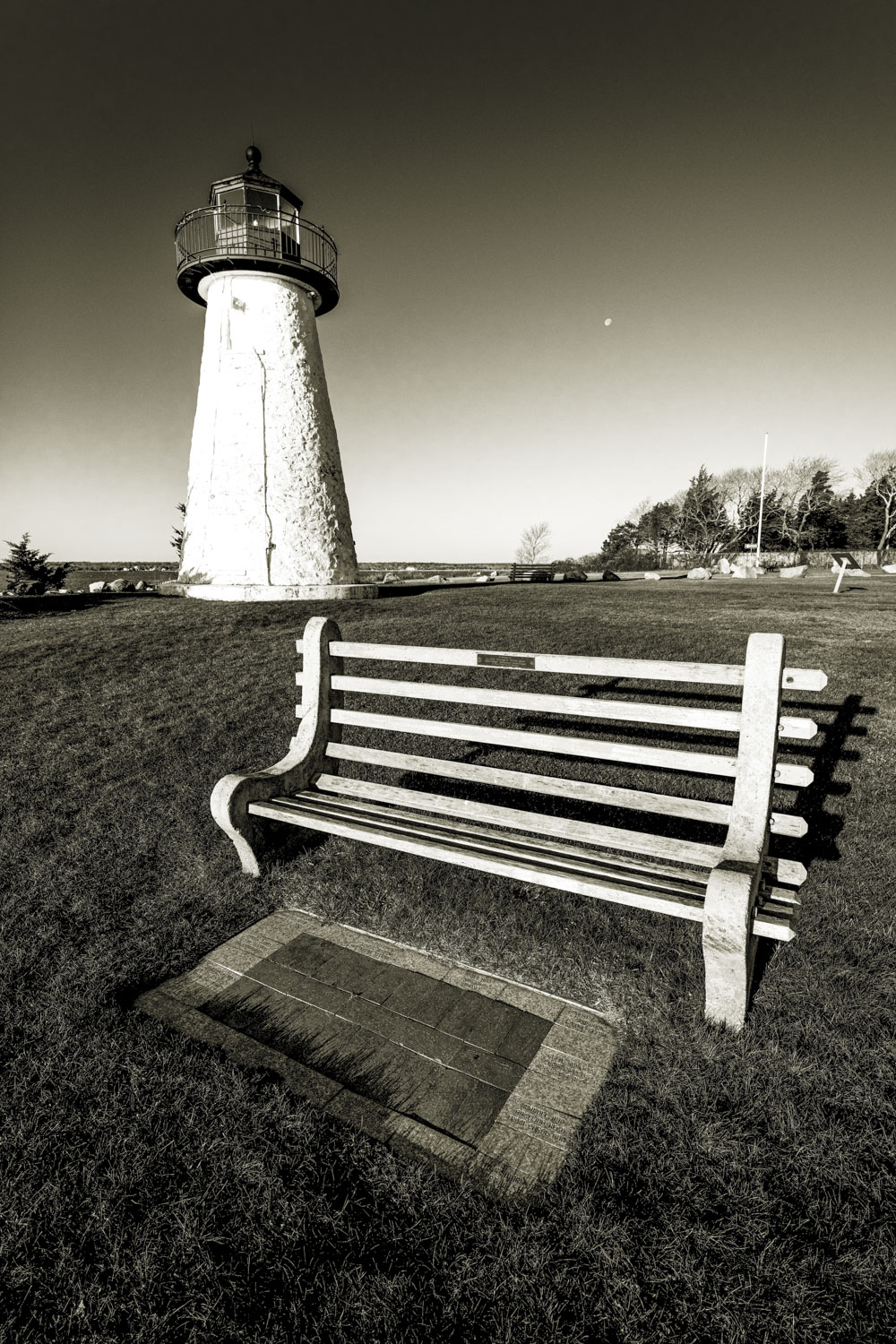 Sepia tone image of a park bench with a lighthouse behind it and the moon in the sky