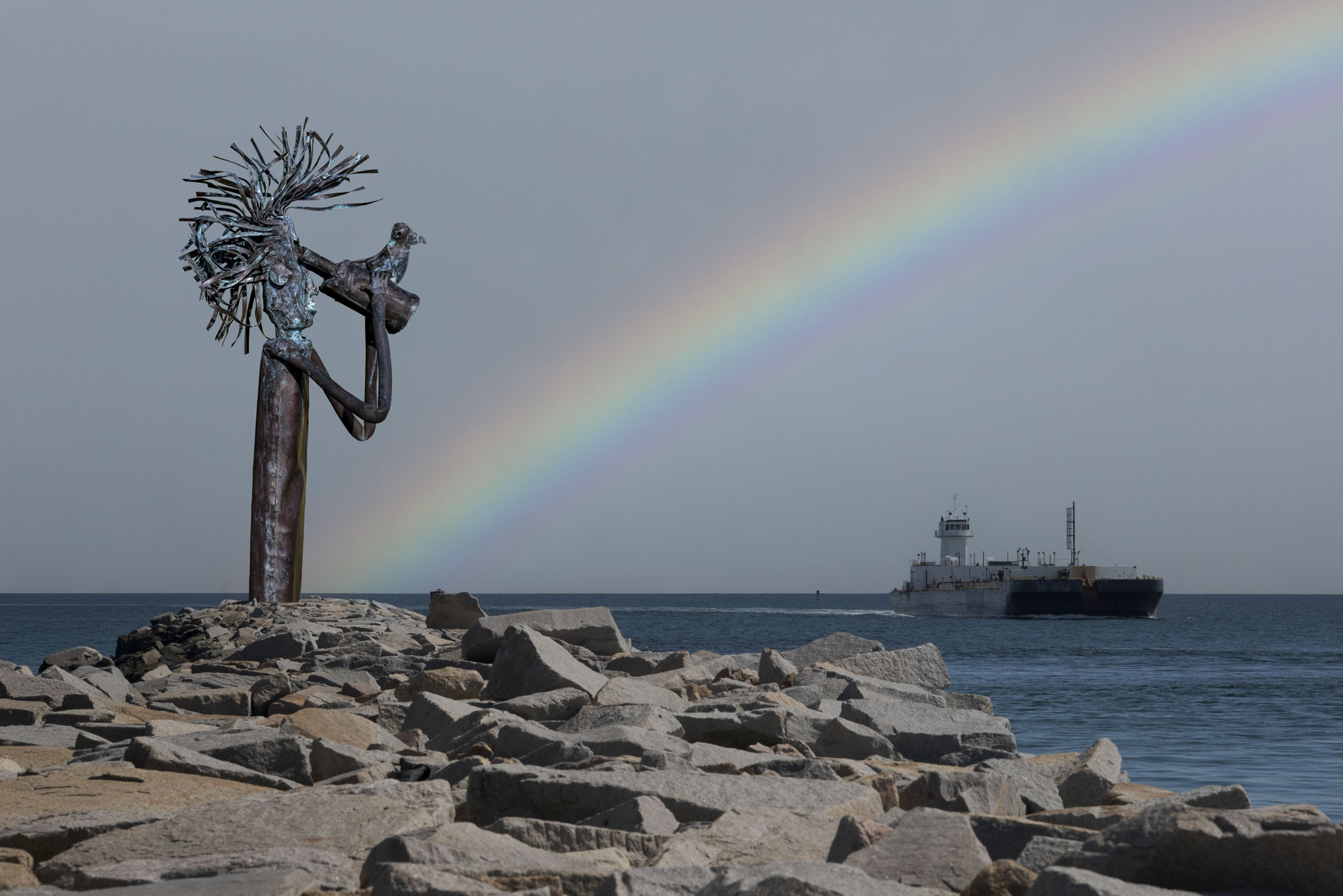 A sculpture with binoculars on rocks, looking out at a barge with a rainbow in the sky