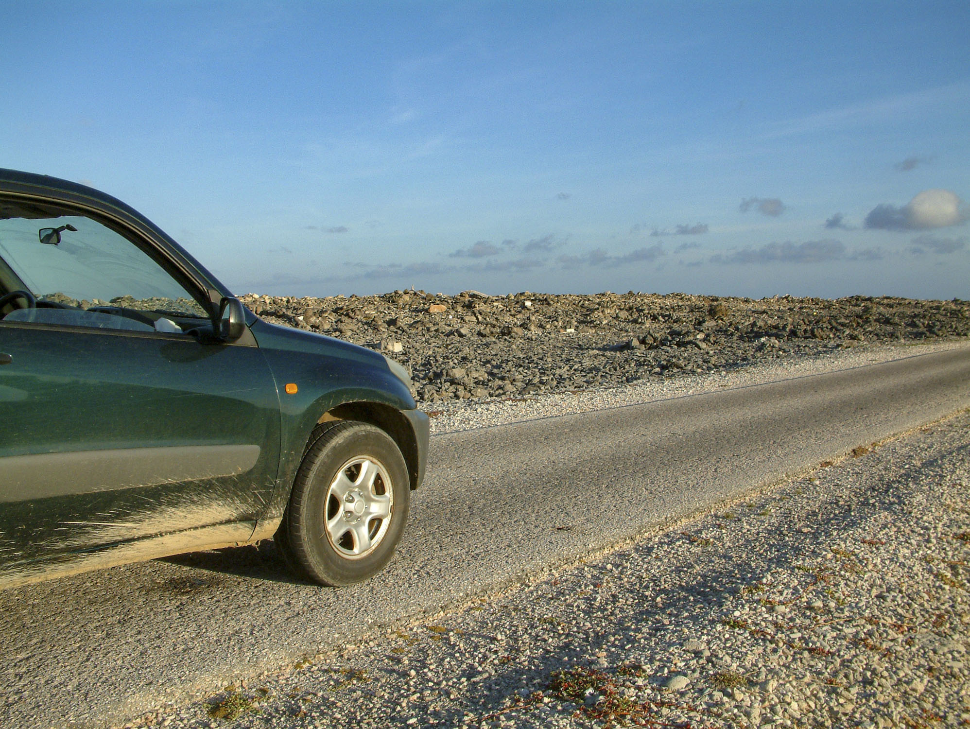 The front of a green car on a flat road in a rocky area